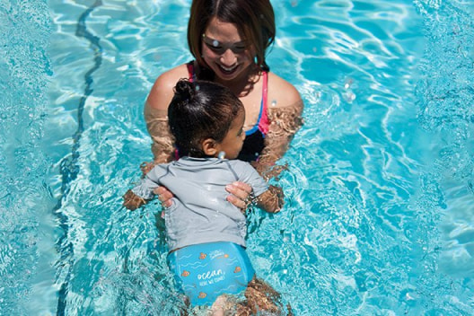 A laughing mother holds her baby wearing a pair of Huggies Little Swimmers Swim Pants in a pool